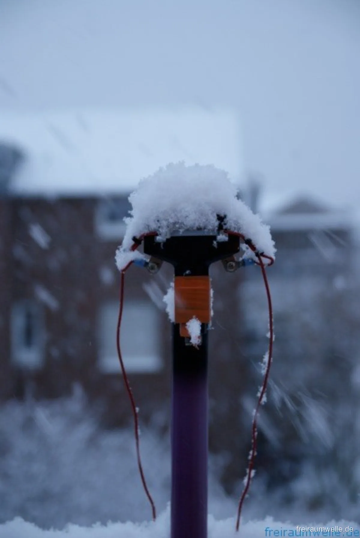 Verschneiter Antennenfußpunkt im Schneetreiben: eine dicke Schneehaube auf dem Kunststoffgehäuse, rote Anschlussdrähte hängen vereist herunter
